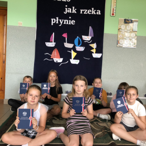  Uczniowie kl. 3 z dyplomami siedzą na dywanie na tle gazetki okolicznościowej.