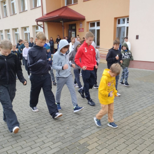 Kacper prezentuje prawidłowy chód sportowy, a uczniowie próbują swoich sił na podwórku szkolnym.